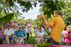 A seated crowd of guests listen attentively to a person speaking and holding up a large stem of bananas.