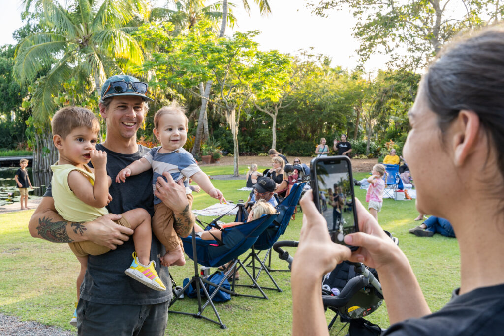 A person poses for a photo while holding two young children. There is another person taking a picture of them with a phone. The background shows a lawn with chairs and blankets on it and people sitting.