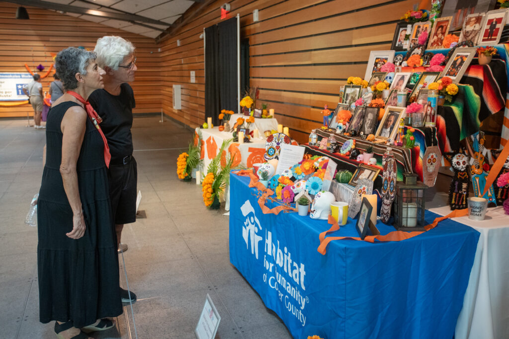 Two guests observe an ofrenda created by the Habitat for Humanity Collier County that has tiered levels featuring photographs of loved ones, candles, marigolds, and calaveras, often referred to as sugar skulls. 