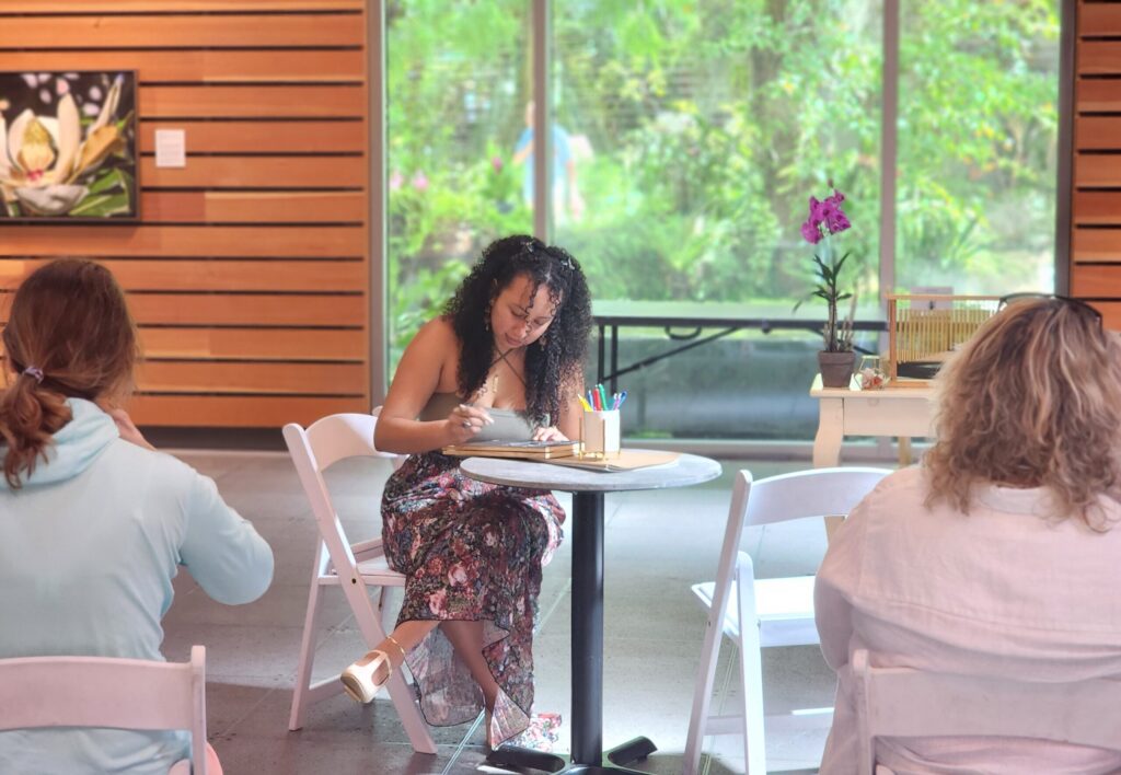 Three people sit at separate tables, two with their back to the camera and the person in the middle facing the camera. We see the person in the middle is writing in a notebook.