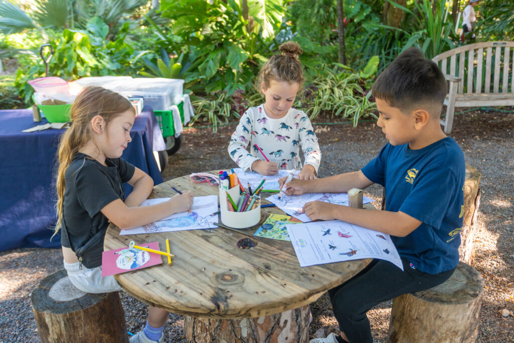 Three children sit outside at a wooden table and chairs made from tree stumps and work on coloring sheets and workbooks. 
