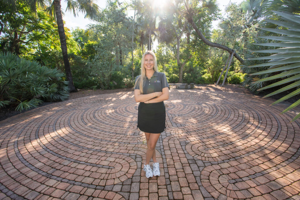 Julia Van Fleet stands within the circular labyrinth, surrounded by dense tropical plants including palm trees and saw palmettos. 