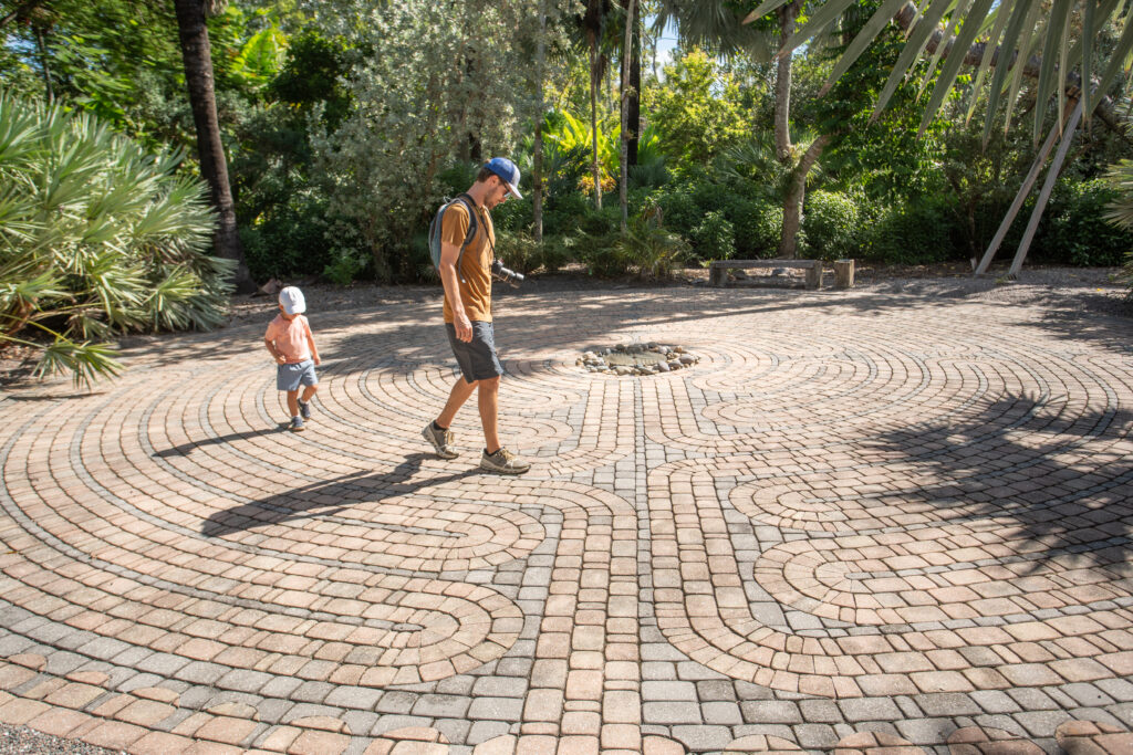 A parent walks the pathway of the labyrinth with their young child following behind them.