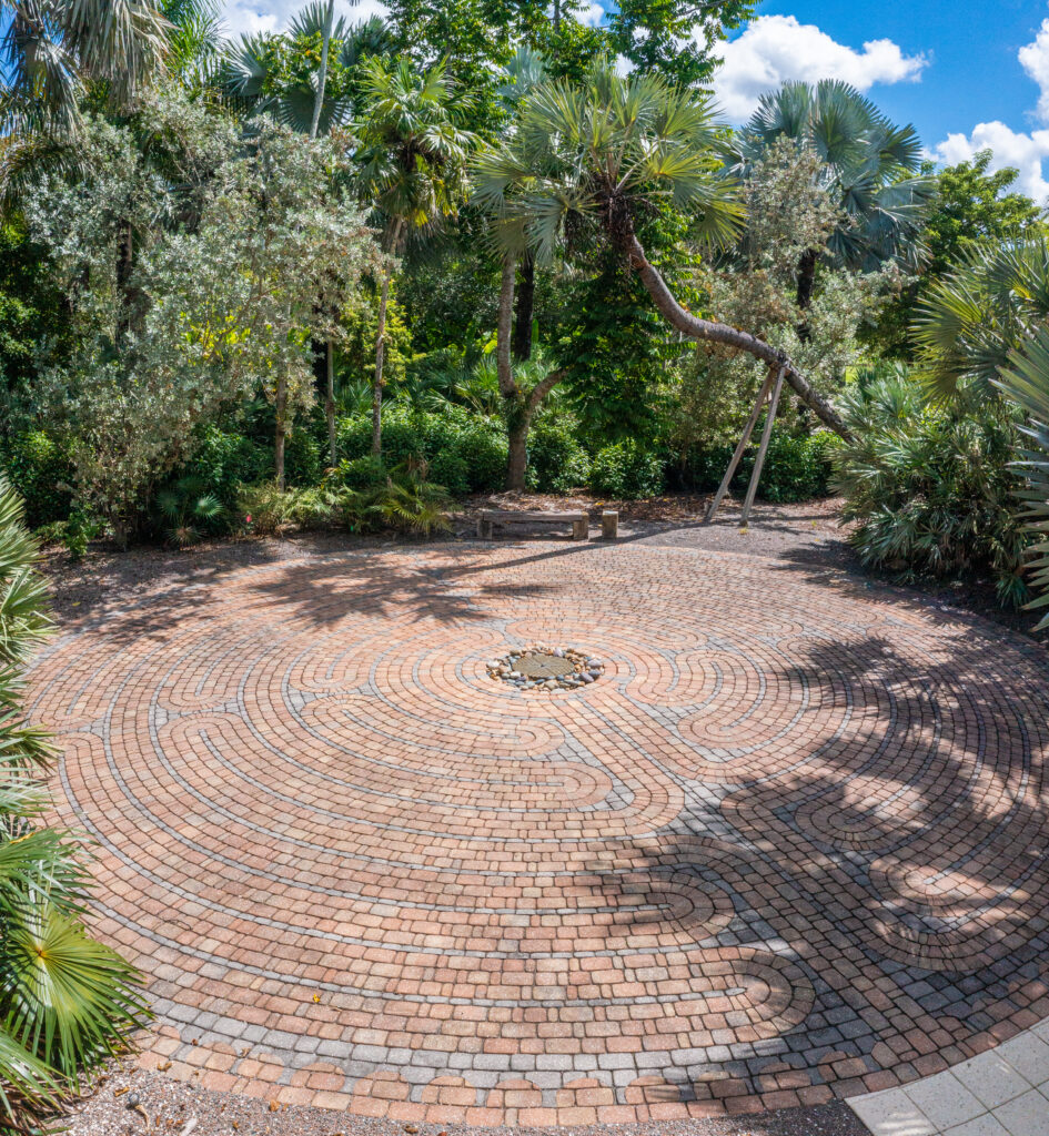 The stone labyrinth is partially shaded by the surrounding foliage. There is a bench tucked away at the edge of the labyrinth in the shade of the buttonwoods and palms. 