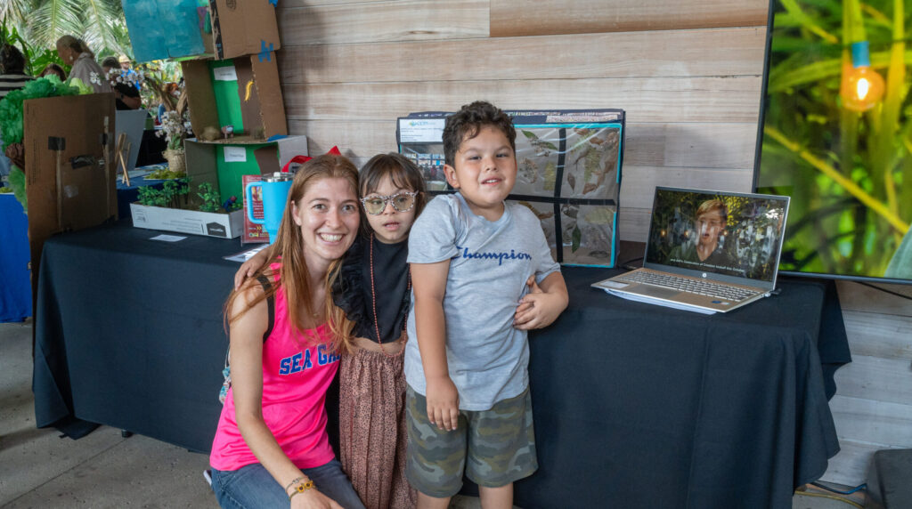 Three people, an adult and two children, pose for a photo in front of a table display.