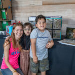 Three people, an adult and two children, pose for a photo in front of a table display.
