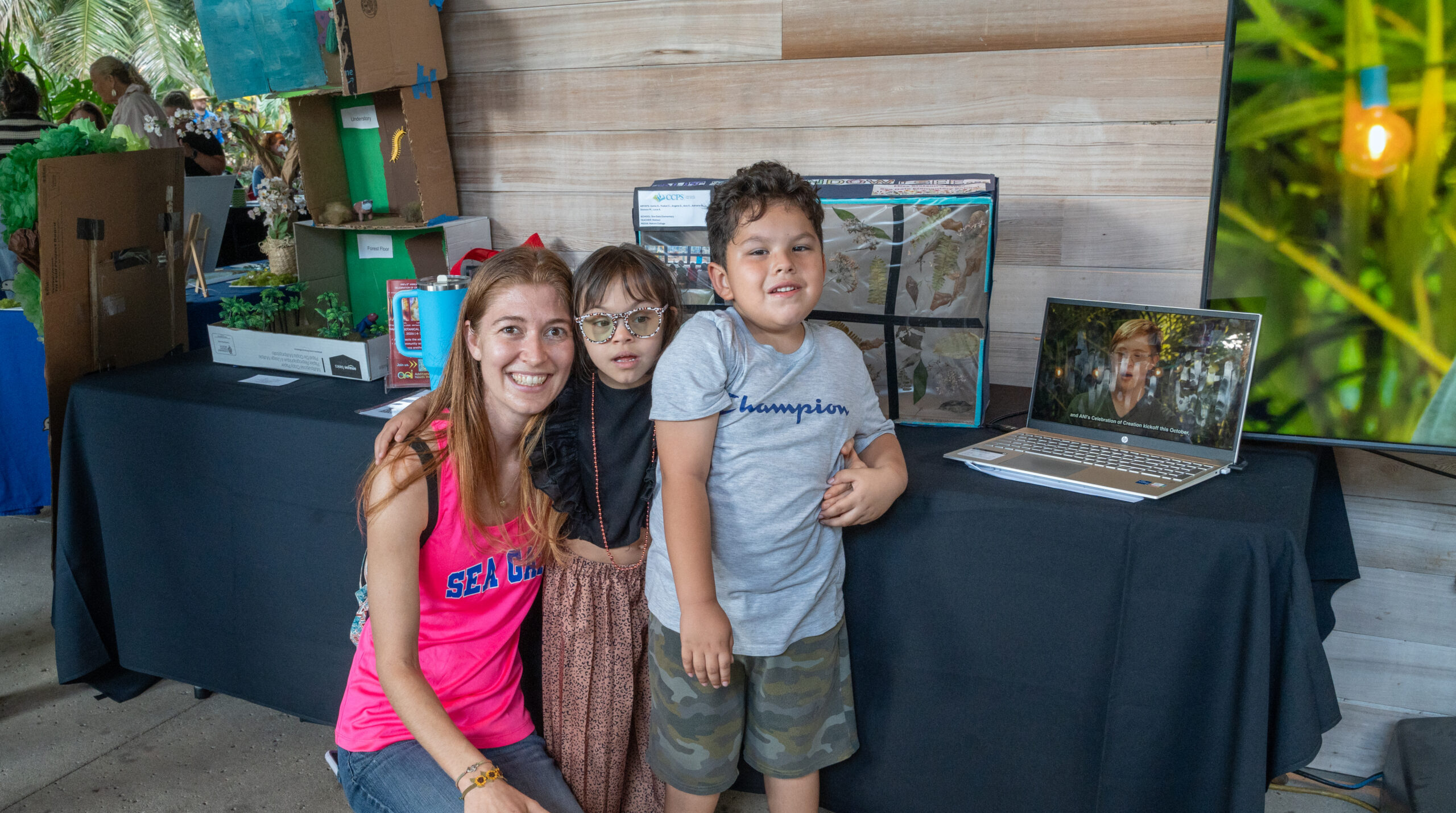 Three people, an adult and two children, pose for a photo in front of a table display.