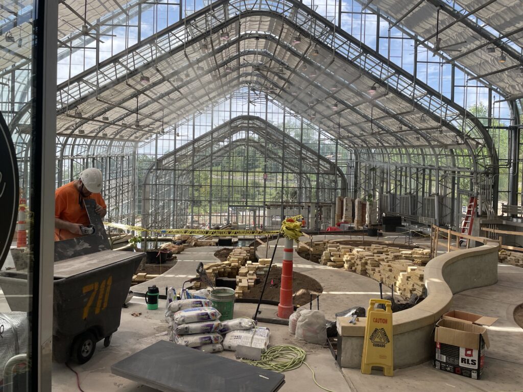 The conservatory is under construction with empty plant beds, piles of rocks, tools, and caution tape marking off pathways. 