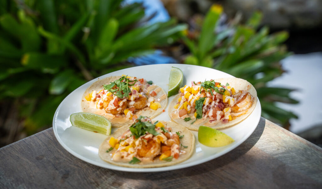 Three shrimp tacos topped with sauce and mango pico de gallo, garnished with lime wedges, sit on a plate on a table. There are green plants in the background.