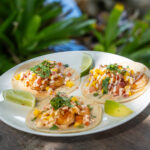Three shrimp tacos topped with sauce and mango pico de gallo, garnished with lime wedges, sit on a plate on a table. There are green plants in the background.