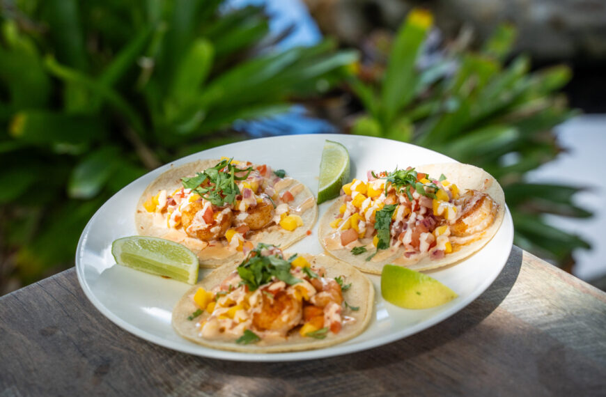Three shrimp tacos topped with sauce and mango pico de gallo, garnished with lime wedges, sit on a plate on a table. There are green plants in the background.