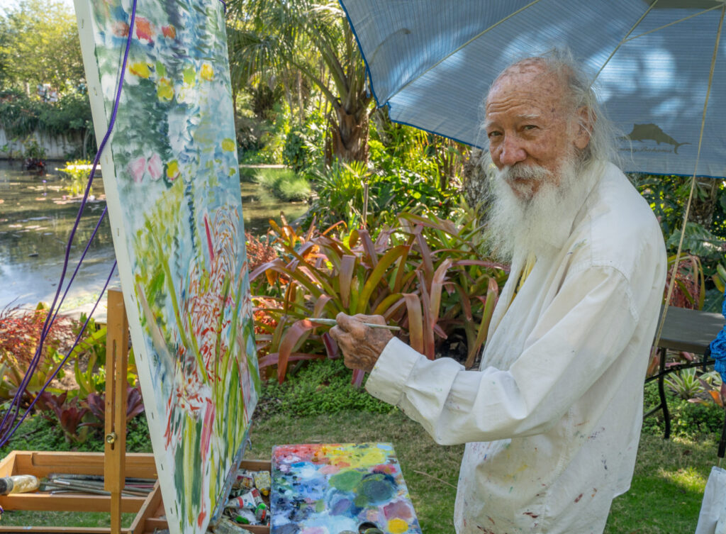 A person stands at an easel with paintbrush in hand. There is a colorful painting in process on the easel and they are surrounded by tropical plants.
