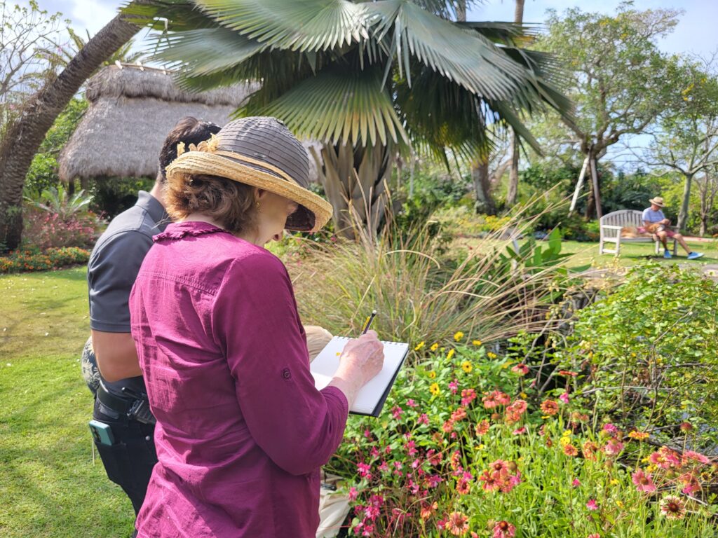 Two people stand and draw in sketchbooks while observing garden foliage. There is a planting of pink and yellow flowers in front of them and a large palm behind them.
