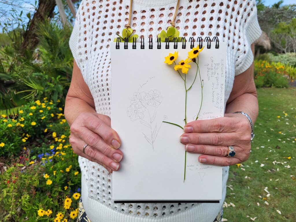 A person holds up a notebook along with three yellow flowers. The paper has sketches and notes about the flowers in the photo.