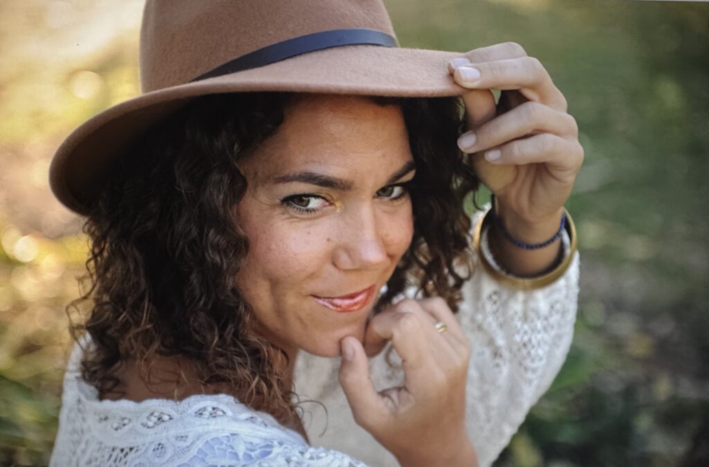 Wearing a brown felt hat, a person poses for a photograph while peering over their shoulder and smiling. 