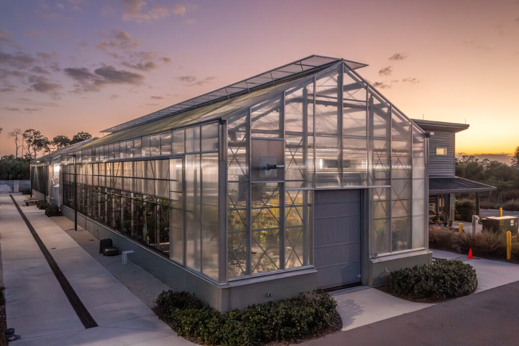 The sunset illuminates a large greenhouse with lights on inside and the shadows of plants within. 