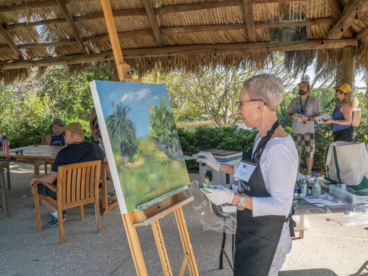 A person stands under a chickee hut and paints a tropical scene on a large canvas on an easel.