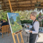 A person stands under a chickee hut and paints a tropical scene on a large canvas on an easel.