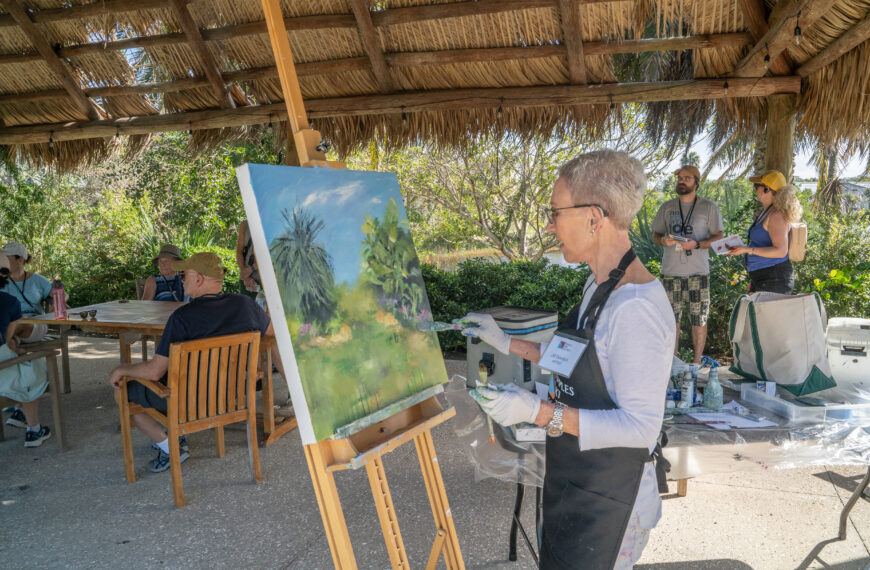 A person stands under a chickee hut and paints a tropical scene on a large canvas on an easel.