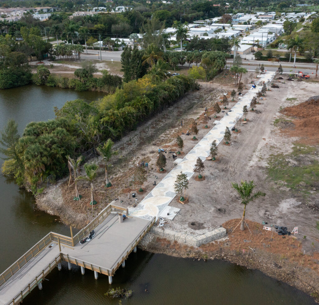Aerial footage shows a straight, concrete walkway lined with new plantings that meets a section of boardwalk going over water.
