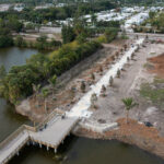 Aerial footage shows a straight, concrete walkway lined with new plantings that meets a section of boardwalk going over water.
