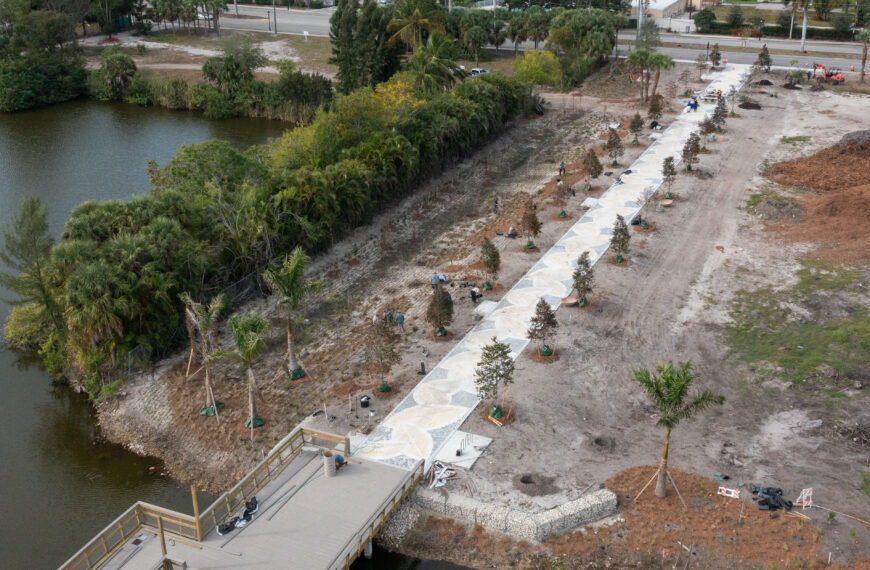 Aerial footage shows a straight, concrete walkway lined with new plantings that meets a section of boardwalk going over water.