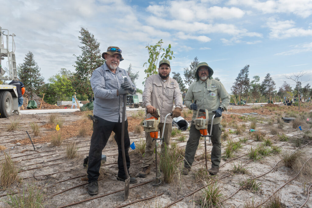 Three Garden employees pose for a photo with their planting tools while standing among rows of newly planted grasses.