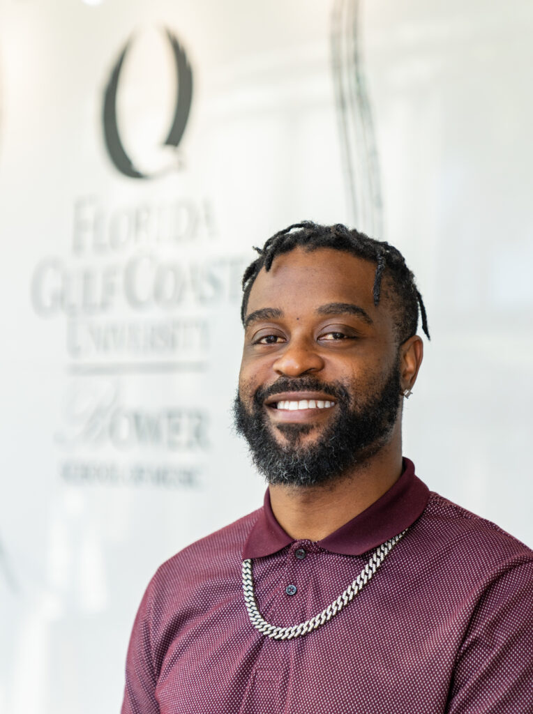 A person wearing a maroon polo shirt with a silver chain necklace smiles for their photo with the Florida Gulf Coast University Bower School of Music logo behind them.