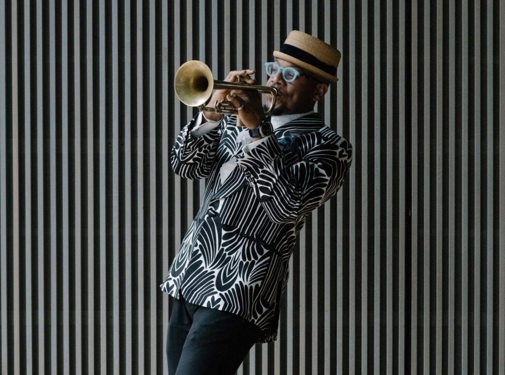 Against a striped black and white backdrop, a person wearing a patterned black and white blazer plays the trumpet.