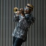 Against a striped black and white backdrop, a person wearing a patterned black and white blazer plays the trumpet.