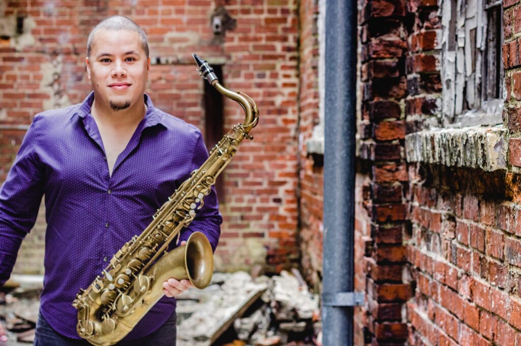 A person, positioned on the left side of the photo, wearing a purple shirt, poses for their photo while holding a saxophone. 