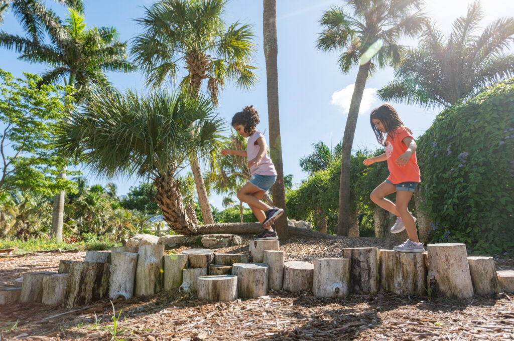 Two children hop across a series of uneven, stationary stumps. 