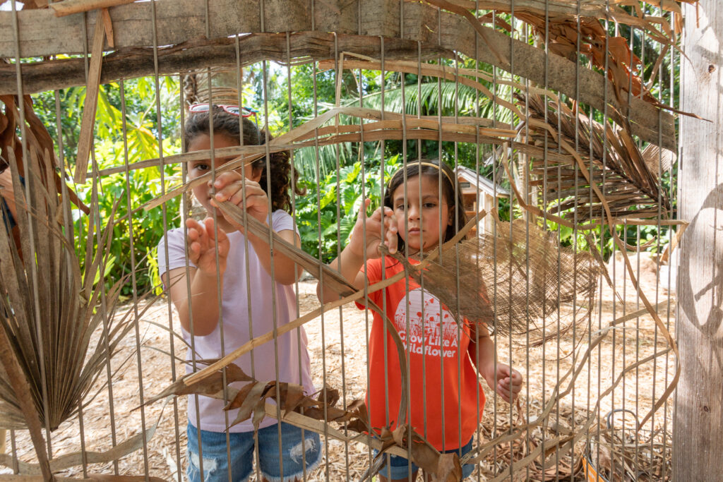 A large weaving loom with dried plant material woven throughout it stands in front of two children who are weaving new pieces onto it. 