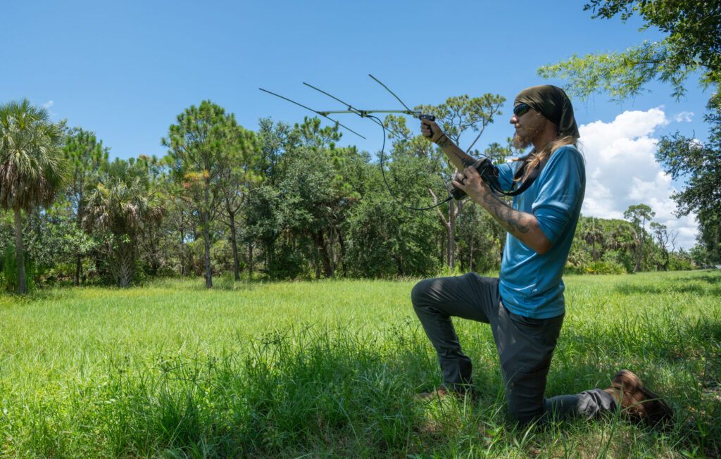 A person kneels on the right side of the photo in a grassy field with trees in the background. The person is holding up an antenna for a radio telemetry device.