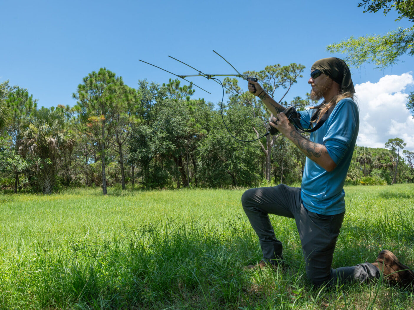 A person kneels on the right side of the photo in a grassy field with trees in the background. The person is holding up an antenna for a radio telemetry device.