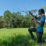 A person kneels on the right side of the photo in a grassy field with trees in the background. The person is holding up an antenna for a radio telemetry device.