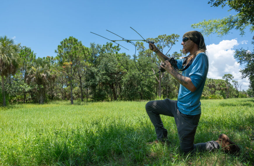 A person kneels on the right side of the photo in a grassy field with trees in the background. The person is holding up an antenna for a radio telemetry device.