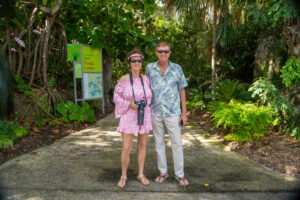 A couple poses for a photo while standing on the sidewalk surrounded by tropical plants. One of them is holding a camera. 