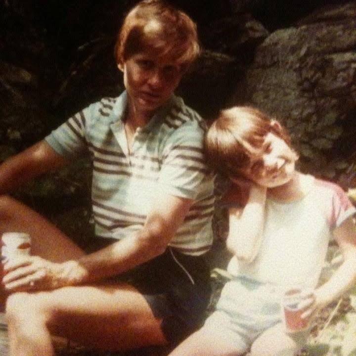 An adult and child pose for a photo while sitting on boulders, each holding a soda can. 