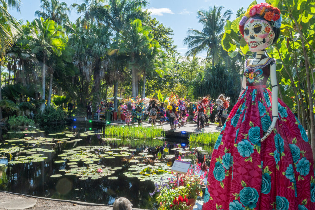 A large Catrina statue in a pink and blue floral dress takes up the right side of the frame and in the background there is a large crowd of people on the far side of a pond.