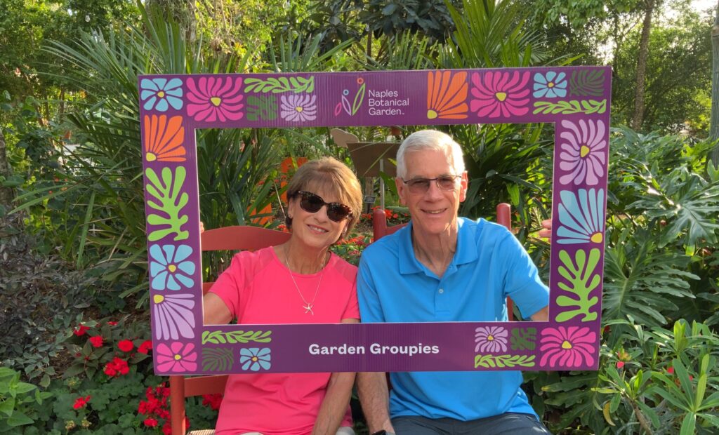 Two people pose for a photo in a tropical garden setting while holding up a frame around them that says Garden Groupies on it and has the Naples Botanical Garden logo. 