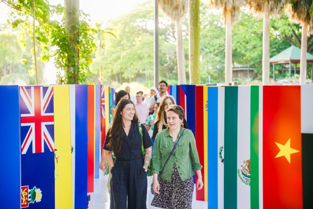 A walkway full of people is lined with columns that represent the flags of the countries from around the world. The sun is shining in the background against tall palm trees.