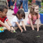 Three children have their hands deep in a garden bed full of dirt.