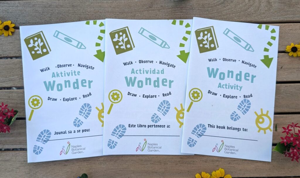 Three white paper booklets are arranged on a wooden deck with small yellow and pink flowers surrounding them. The booklets have the same Wonder Activity title and design, but in three separate languages: Haitian Creole, Spanish, and English. 