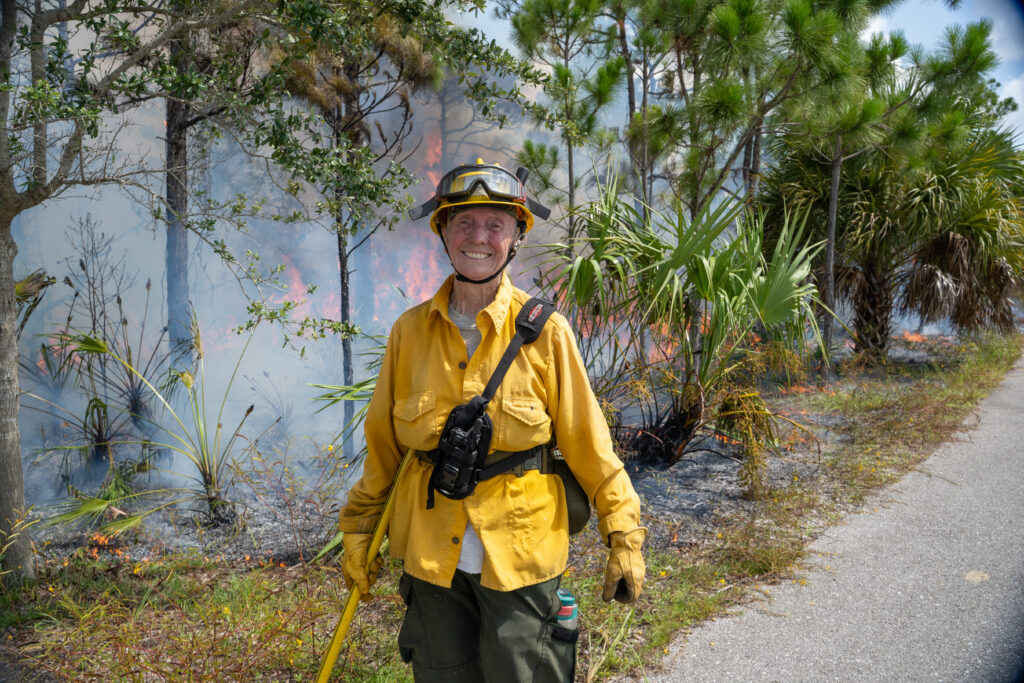 A person smiles for the camera in their fire protective gear while the land behind them burns and smolders. There are pine trees, palms, and shrubs. 