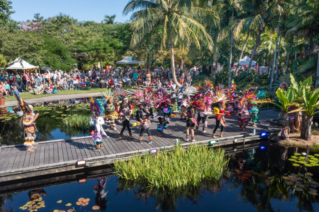 A large crowd of onlookers watch from the background while dancers in bright, colorful outfits dance on a stage that spans a water garden.