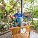 A Garden educator in a blue shirt stands behind a rolling cart with a tropical garden bed behind them. The tabletop has large props on it such as a large flower model, a spinning wheel, and a globe.