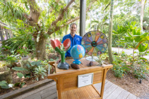 A Garden educator in a blue shirt stands behind a rolling cart with a tropical garden bed behind them. The tabletop has large props on it such as a large flower model, a spinning wheel, and a globe.