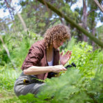 A person kneels down photograph a plant up close. They are surrounded by ferns, shrubs, and other native plants.