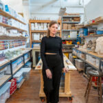 A person poses for a photo while leaning against a table in an art studio. The walls and counter tops on either side of them are full of supplies.